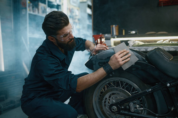 Jongeman werknemer draagt beschermende bril schuren motorfiets van oude verf tijdens het werk bij reparatieservice - Foto, afbeelding