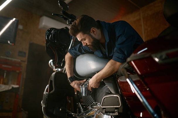 Male mechanic working in garage and repairing custom motorcycle. Creative authentic workshop concept - 写真・画像