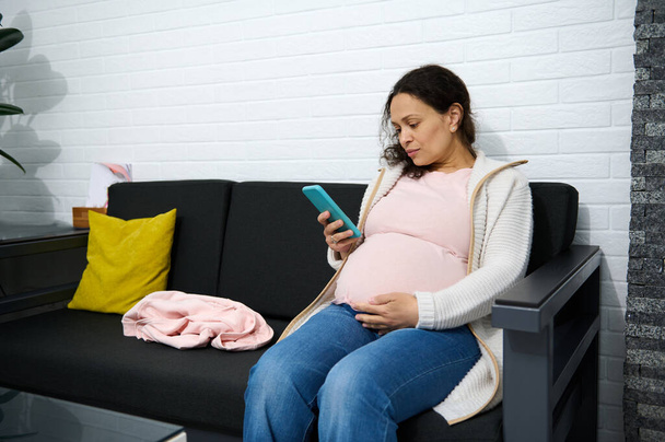 Monietninen aikuinen raskaana oleva nainen tarkistaa matkapuhelimensa istuessaan sohvalla gynekologisen lääkäriaseman odotussalissa. - Valokuva, kuva