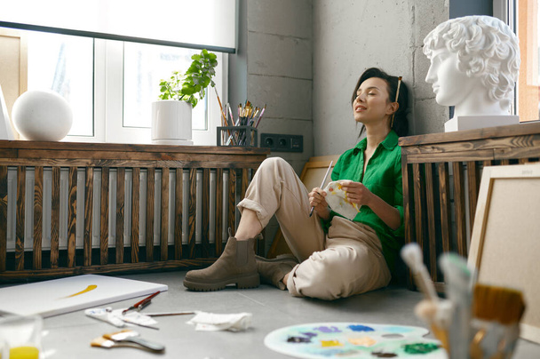 Relaksująca się młoda artystka siedząca na podłodze przed rysunkiem akcesoria do przerwy i odpoczynku z zamkniętymi oczami. Kreatywna zabawa hobby i poszukiwanie inspiracji koncepcja - Zdjęcie, obraz