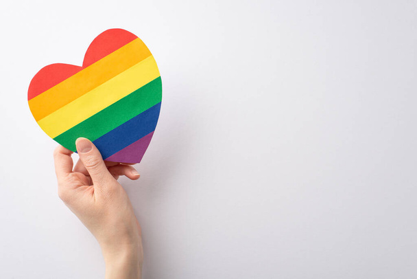 Feiern Sie LGBTQ History Month mit diesem Foto einer weiblichen Hand, die eine herzförmige regenbogenfarbene Karte auf weißem Hintergrund mit Platz für Text oder Werbung hält - Foto, Bild