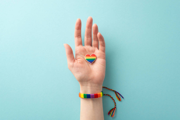 Honra el Mes de la Historia LGBTQ con esta foto en primera persona de una mano femenina que lleva una pulsera de color arco iris y sostiene una insignia de pin en forma de corazón sobre un fondo azul pastel - Foto, imagen