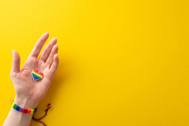 Observeer LGBTQ Geschiedenis Maand met deze eerste persoon bovenaanzicht foto van vrouwelijke hand dragen van een regenboog-gekleurde armband en met een hart-vormige pin badge op gele achtergrond met ruimte voor tekst of reclame - Foto, afbeelding