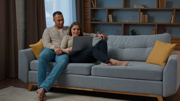Diverse multiracial couple sitting on sofa surfing online website on laptop using computer at home relaxing woman and man talking buying retail goods shopping online make order spend weekend on couch - Zdjęcie, obraz