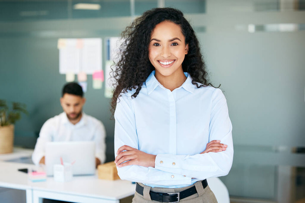 Driven and passionate. an attractive young businesswoman standing in the office with her arms folded while her colleague works behind her - Foto, afbeelding
