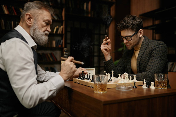 Rodzina inteligentnych ludzi grająca w szachy, paląca cygara i pijąca whisky. Starsi i młodsi mężczyźni siedzą przy stole i patrzą na szachownicę. Weekendowy wieczór w domu koncepcja aktywności męskiej - Zdjęcie, obraz