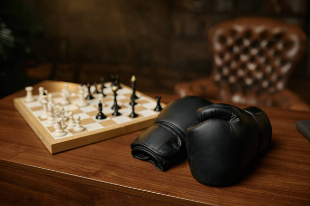 Houten tafel in de kamer met zwarte leren bokshandschoenen en schaakbord met schaakstukken. Schaakconflict concept - Foto, afbeelding