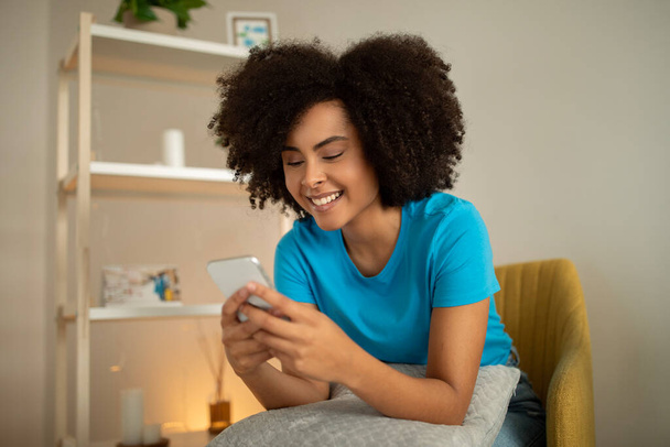 Aplicación para la comunicación. Sonriente milenaria rizado mujer negra escribiendo en el teléfono inteligente, ver vídeo, leer el mensaje en el interior de la sala de estar. Gadget y redes sociales, blog y chat de forma remota en casa - Foto, imagen