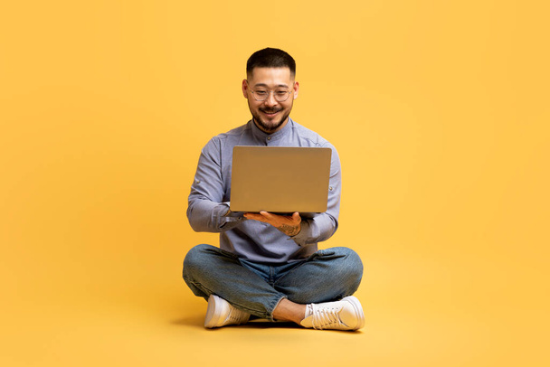 Glücklicher asiatischer Mann mit Laptop, während er auf dem Boden über gelbem Studiohintergrund sitzt, erfolgreicher Millennial Male Freelancer oder Blogger Guy, der auf dem Computer im Internet surft und Remote Business genießt - Foto, Bild