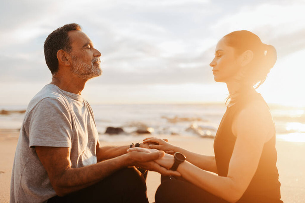 Happy calm mature caucasian man and woman in sportswear sit, practice yoga, meditate in morning on beach, outdoor. Couple enjoy workout together, peace, health care, body care in summer - Photo, Image