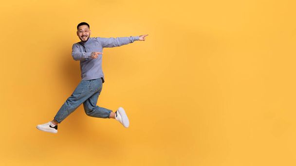 Look At This. Cherful asian man pointing aside while jumping up in air, positive young millennial male having fun and demonstrating free copy space on yellow studio background, panorama - Photo, Image