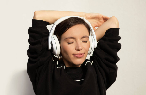 Smiling young caucasian lady with headphones enjoys music, rests from workout with close eyes on white wall background, close up. Sports, fitness and audio app, weight loss, health care with music - Photo, Image