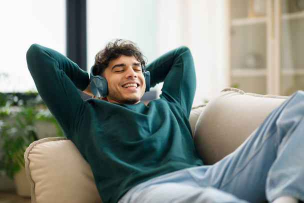 Bliskowschodni Młody człowiek relaksujący się nosząc słuchawki i słuchając muzyki, leżąc na wygodnej kanapie trzymając za ręce w domu. Facet dobrze się bawiący, cieszący się ulubioną piosenką. Wypoczynek weekendowy - Zdjęcie, obraz