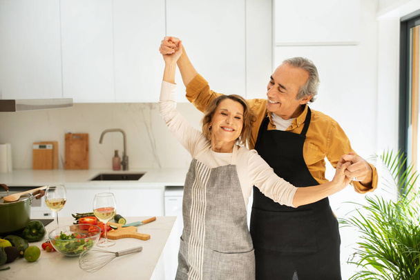 Αγαπώντας το ζευγάρι τελειόφοιτων που χορεύουν ενώ μαγειρεύουν μαζί στην κουζίνα, ευτυχισμένος σύζυγος που απολαμβάνει το χρόνο με την όμορφη σύζυγό του, αντιγράφει το χώρο. Σχέση, γάμος, συνταξιοδότηση - Φωτογραφία, εικόνα