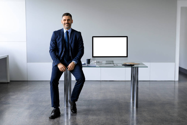 Resmi takım elbiseli yakışıklı orta yaşlı bir işadamı boş bilgisayar monitörüyle ofis masasının yanında oturuyor ve tasarımın için bedava alan sunuyor. İş reklamı konsepti - Fotoğraf, Görsel