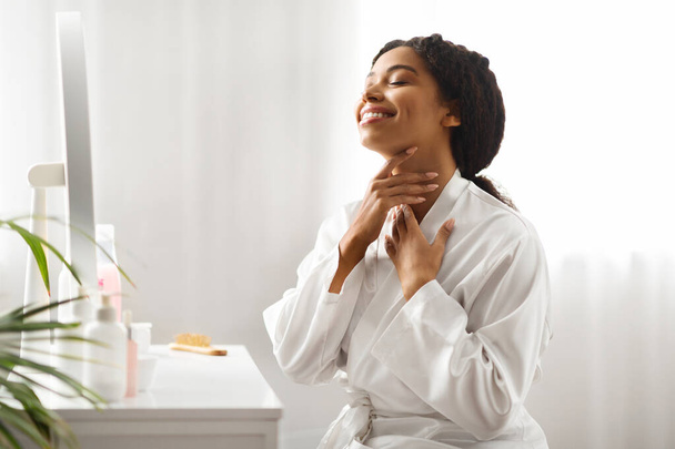 Hermosa mujer negra tocándose el cuello, haciendo masaje de levantamiento de la piel en casa, sonriendo feliz joven afroamericana mujer usando túnica de seda blanca sentada en el tocador en el dormitorio, espacio de copia - Foto, imagen