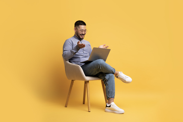 Cherful junger asiatischer Mann macht Videoanruf auf Laptop, während er im Sessel über gelbem Studiohintergrund sitzt, aufgeregter Millennial Guy schaut auf Computerbildschirm und spricht auf Webkamera, Kopierraum - Foto, Bild