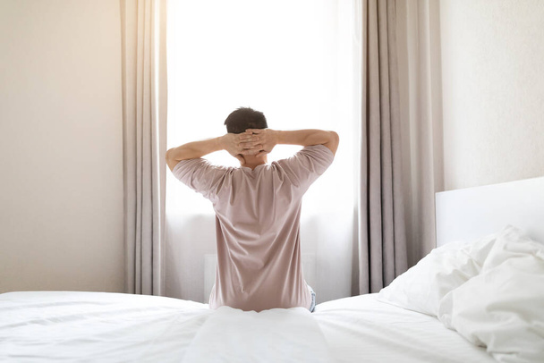 Back view of full-rested brunette man wearing pajamas sitting on bed and stretching body after waking up in the morning, looking at window, sun flare, copy space. Comfortable healthy sleep concept - Zdjęcie, obraz