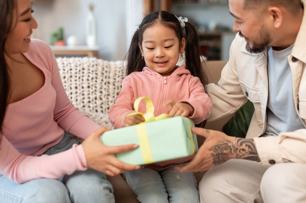 Czas na prezenty. Wesoła japońska rodzina świętuje urodziny córek dziecka, dając opakowane pudełko z prezentami do cute dziecko siedzi na kanapie w domu. Koncepcja świętowania świąt. Przycięte, selektywne ogniskowanie - Zdjęcie, obraz