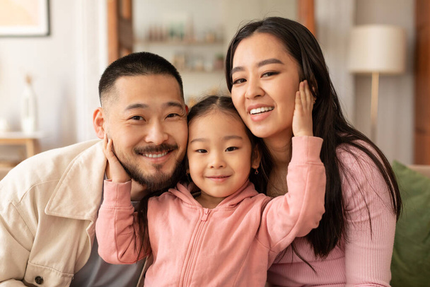Unité familiale. Portrait d'une belle famille coréenne de trois embrassant exprimant des émotions positives posant à la maison. Les parents étreignant leur adorable fille enfant souriant à la caméra ensemble - Photo, image