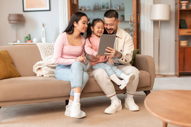 Family Online Lazer. Pais coreanos e pequena filha usando Tablet assistindo desenhos animados on-line no computador sentados juntos na sala de estar moderna interior. Conceito de Internet e Diversão - Foto, Imagem