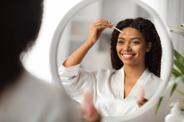 Conceito de Cuidados com a pele. Mulher negra bonita aplicando soro facial perto do espelho, mulher afro-americana feliz fazendo rotina diária de cuidados com a pele em casa, testando novos cosméticos, foco seletivo na reflexão - Foto, Imagem
