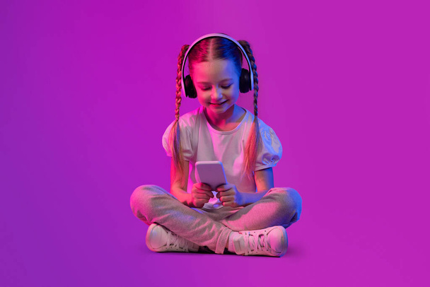 Ψηφιακή Native, Generation Alpha Concept. Preteen παιδί ευτυχισμένη αρκετά αξιολάτρευτο κορίτσι στο καλοκαιρινό ντύσιμο κάθεται στο πάτωμα πάνω φουτουριστικό φόντο, χρησιμοποιώντας ασύρματα ακουστικά και smartphone, αντίγραφο χώρο - Φωτογραφία, εικόνα