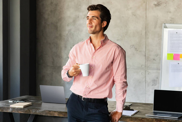 Successful european businessman handsome young man in formal outwear standing by desk, holding mug. CEO manager, leader have coffee break at office, looking at copy space - Foto, immagini