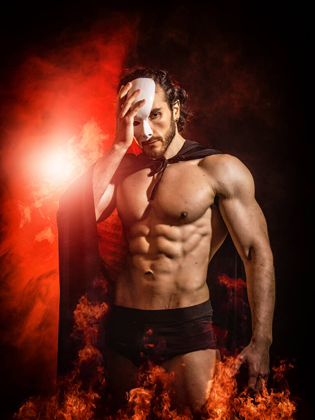 Shirtless muscular male with naked torso in cloak and underpants standing in darkness, among fire and covering part of face with white mask - Foto, afbeelding