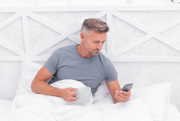 Happy guy use mobile phone while relaxing in bed with morning coffee ot tea, sms. - Φωτογραφία, εικόνα