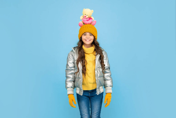 happy kid in puffer jacket and hat. childhood. teen girl hold toy bear. child wear warm clothes on blue background. express positive emotion. winter fashion. love toy for valentines day. - Фото, зображення