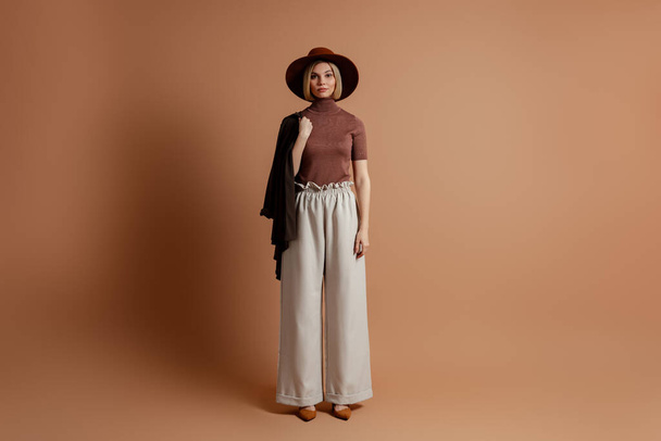 Full length of fashionable young woman in elegant hat standing against brown background - Foto, Bild
