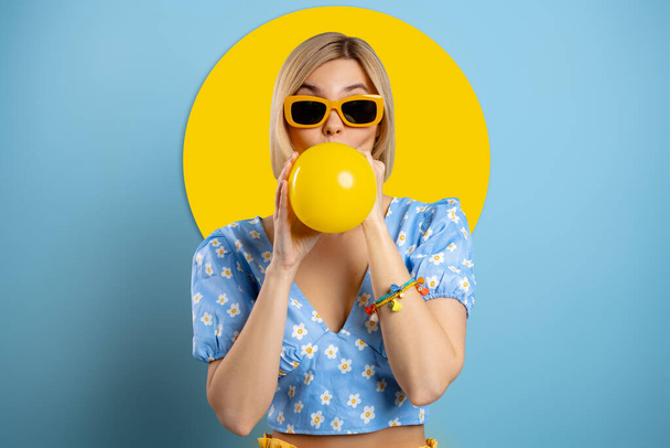 Playful young woman blowing up a balloon while standing against blue background - Fotografie, Obrázek