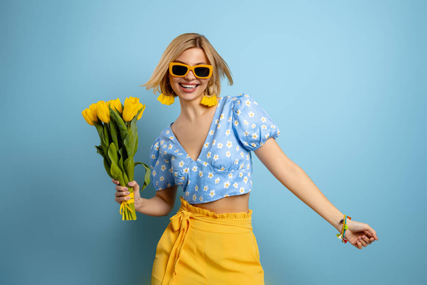 Attractive young woman holding bunch of tulips and dancing against blue background - Foto, Imagen