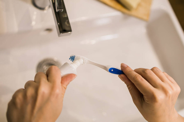 Female hand holding toothbrush with toothpaste applied on it in bathroom. Close up of female hand ready for brushing teeth. Young woman hand holding toothbrush with white tooth paste - Foto, Imagem