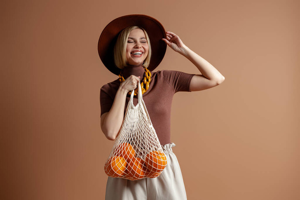 Happy young woman in elegant hat carrying mesh bag with oranges against brown background - Fotó, kép