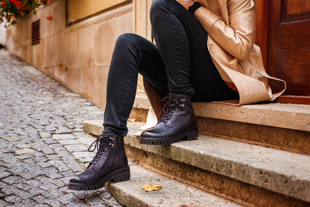 Trençkot giyen ve ayak bileği çizmeli bir kadın şehirde merdivenlerde oturuyor. Modaya uygun siyah deri ayakkabı. Sokak modası - Fotoğraf, Görsel