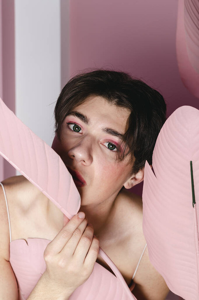 Close-up Portret van een tedere homo in een roze jurk poseren met een geschilderde tropische plant of bladeren. Glamour homoseksuele jonge man met make-up poseren in een meisjesachtige outfit. hoofdschot. - Foto, afbeelding