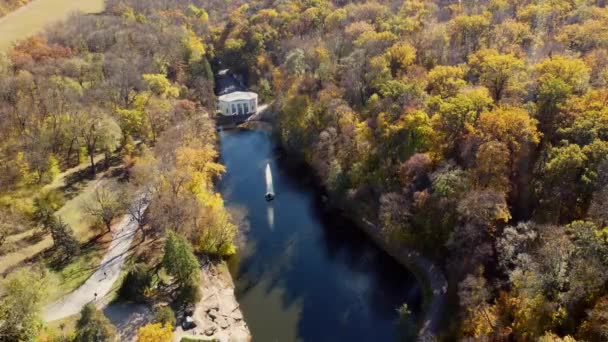 Many trees with yellow leaves, lake with fountain in center, architecture, big stones, paths walkways, people walking on paths on sunny autumn day. Aerial drone view. Panoramic autumn landscape park - Footage, Video