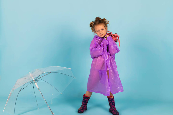 Urocza dziewczynka w fioletowym płaszczu przeciwdeszczowym i gumowych butach trzyma czysty parasol. Dziecko ubrane na deszczową pogodę. Izolacja na niebieskim tle. - Zdjęcie, obraz