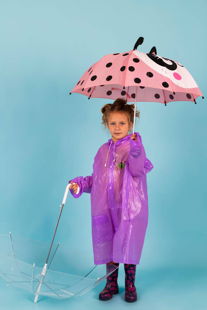 Το χαριτωμένο κοριτσάκι με το μωβ αδιάβροχο και τις λαστιχένιες μπότες κρατάει δύο ομπρέλες. Ένα παιδί ντυμένο για βροχερό καιρό. Απομονωμένα σε μπλε φόντο. - Φωτογραφία, εικόνα