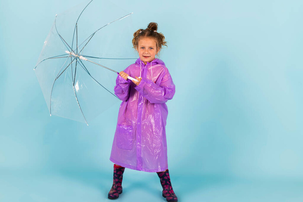 Charming little girl in a purple raincoat and Rubber boots holds a clear umbrella. A child dressed for a rainy weather. Isolated on blue background. - Foto, Bild