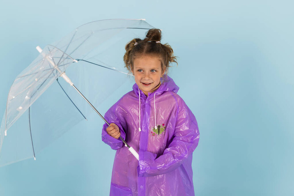 Mor yağmurluklu ve lastik çizmeli sevimli küçük bir kız şemsiye tutuyor. Yağmurlu bir hava için giyinmiş bir çocuk. Mavi arkaplanda izole. - Fotoğraf, Görsel