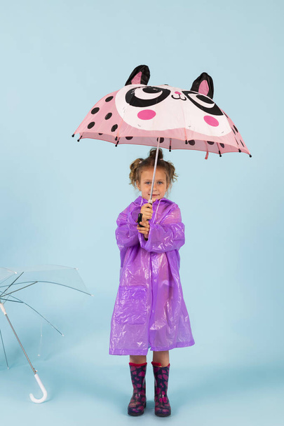 Okouzlující holčička v purpurovém plášti a gumových botách drží deštník. Dítě oblečené na deštivé počasí. Izolováno na modrém pozadí. - Fotografie, Obrázek