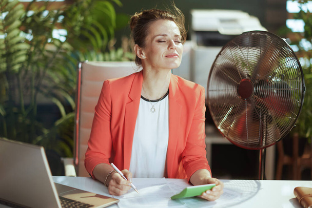 Sustainable workplace. smiling modern 40 years old accountant woman in modern green office in red jacket with calculator, electric fan and laptop. - Foto, imagen