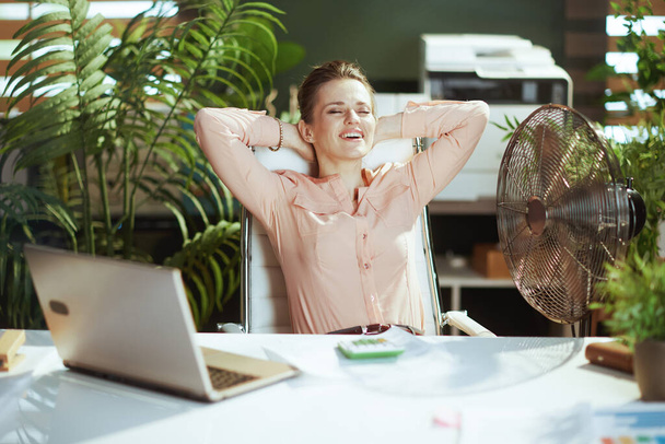 Sustainable workplace. happy modern middle aged bookkeeper woman at work with electric fan and laptop. - Foto, Bild