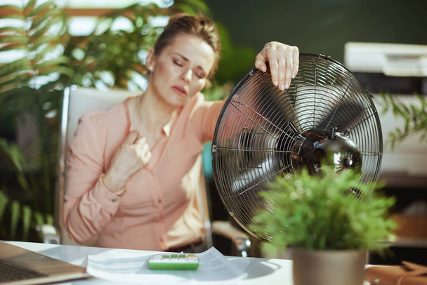 Sustainable workplace. modern 40 years old woman worker in modern green office with electric fan suffering from summer heat. - Foto, Imagem