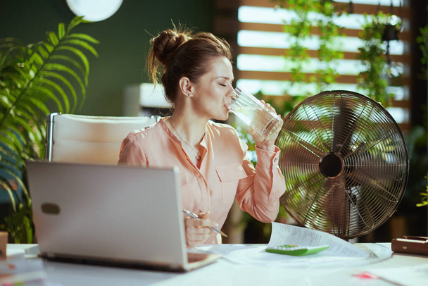 Sustainable workplace. smiling modern 40 years old woman employee at work with electric fan, cup of water and lemon. - 写真・画像