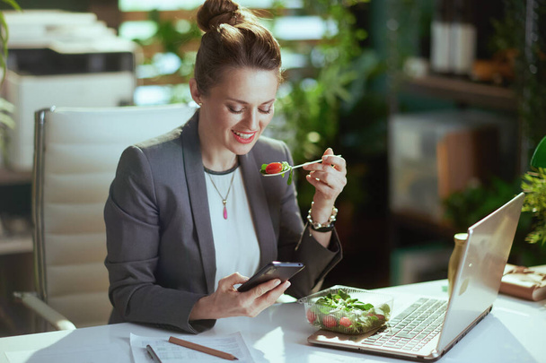 Sustainable workplace. happy modern 40 years old business woman in a grey business suit in modern green office with laptop eating salad and using smartphone applications. - Foto, afbeelding