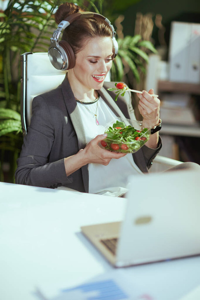 Sustainable workplace. smiling modern female employee in a grey business suit in modern green office with laptop and headphones eating salad. - Valokuva, kuva